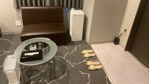 A small room with a brown sofa, a glass table with a remote control, a white appliance, and a pair of slippers on the floor. - My Dear - Adults Only (Osaka)