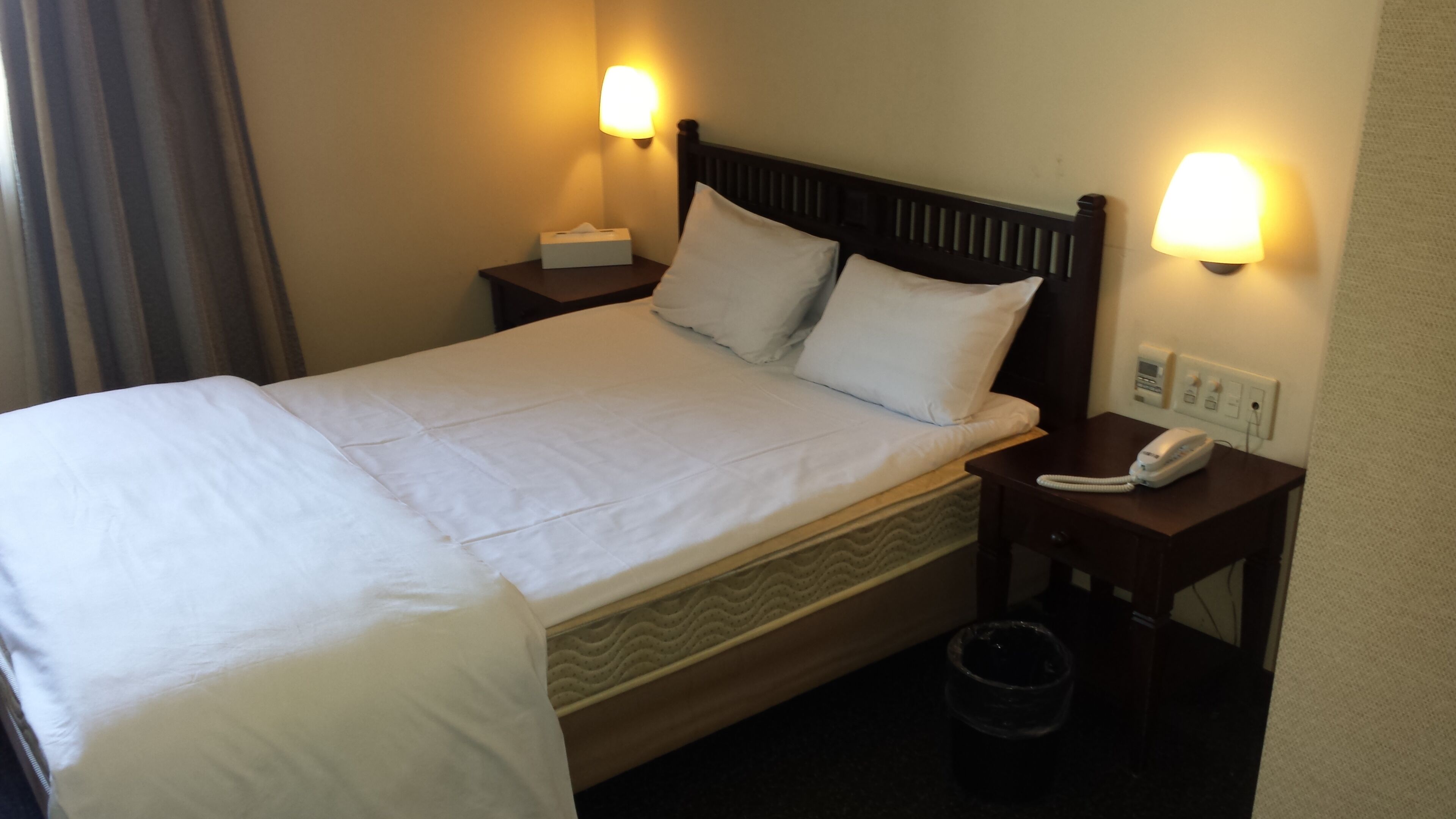 A hotel room with a bed, two bedside lamps, a telephone, and a waste bin.