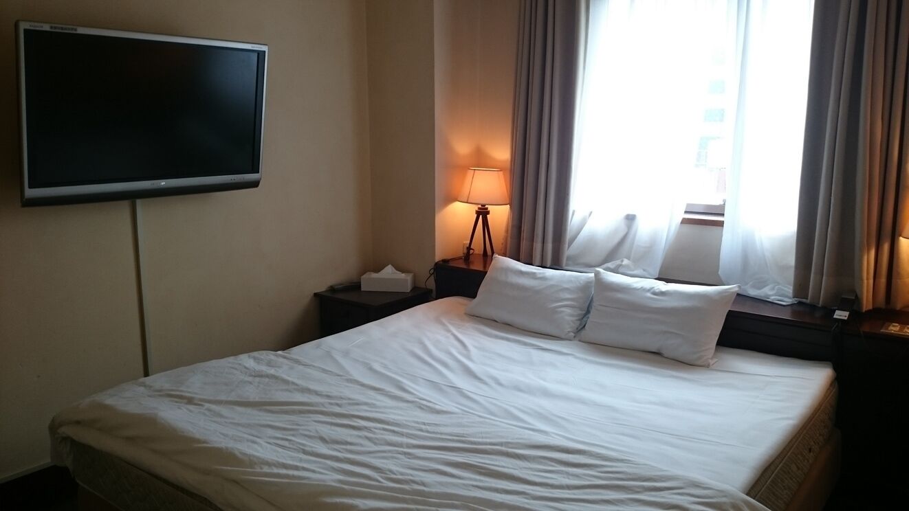Une chambre d’hôtel avec un lit, un téléviseur à écran plat fixé au mur, une table de chevet avec une lampe et une fenêtre avec des rideaux.