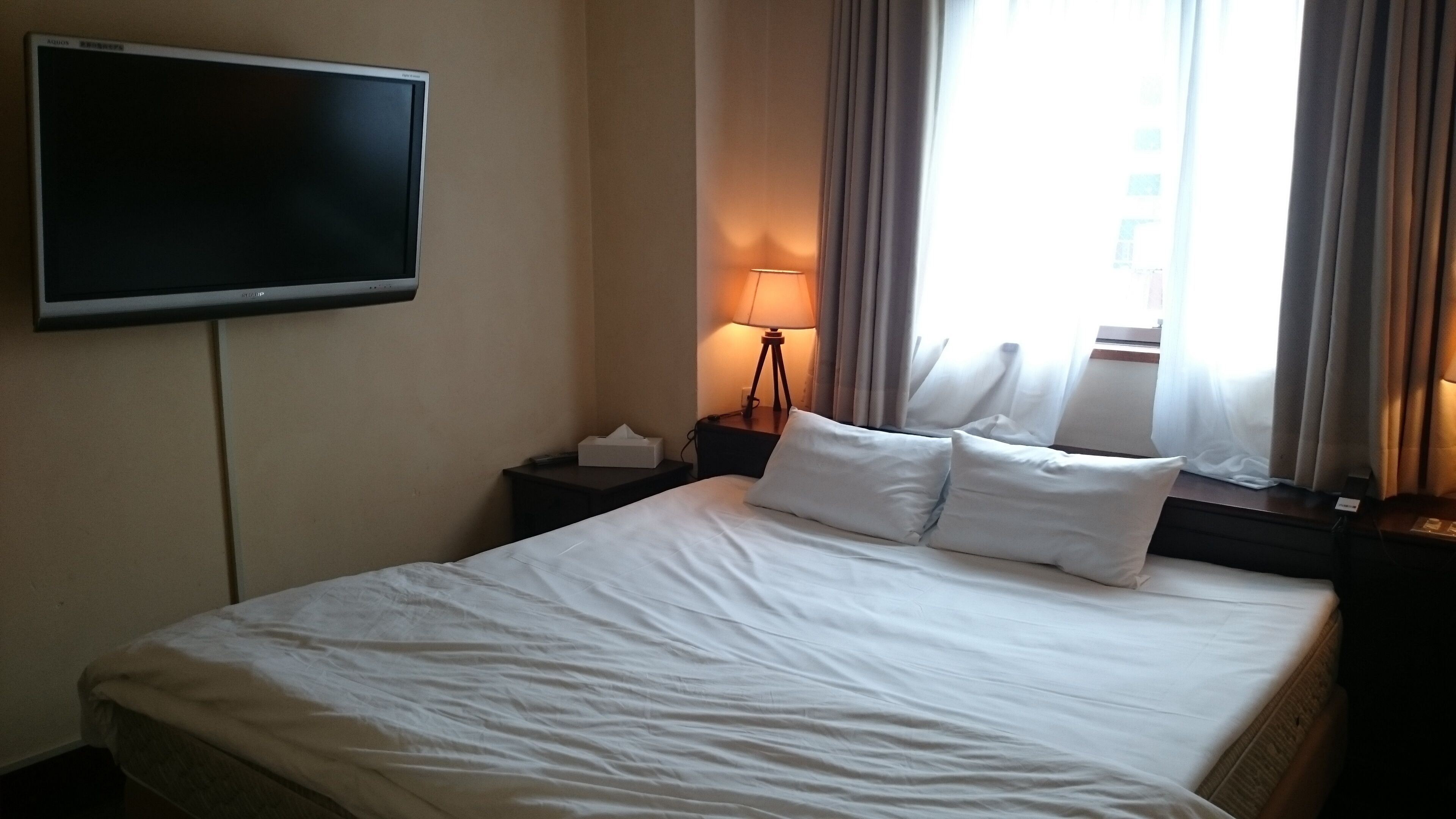 A hotel room with a bed, a TV mounted on the wall, a bedside lamp, and a window with curtains.
