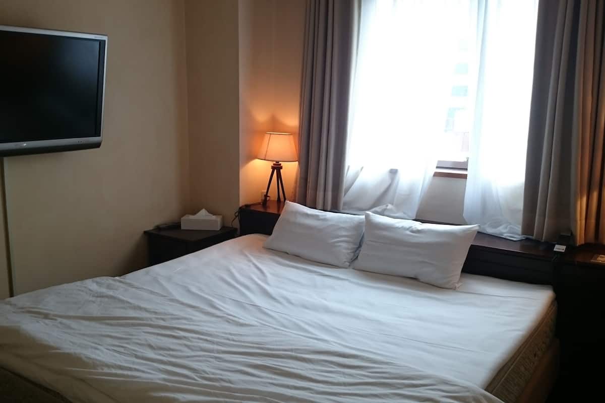 A hotel room with a bed, a TV mounted on the wall, a bedside lamp, and a window with curtains.