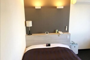 Una habitación de hotel con una cama, una lámpara de noche, una mesita con un teléfono y un cubo de basura, y dos luces en la pared.