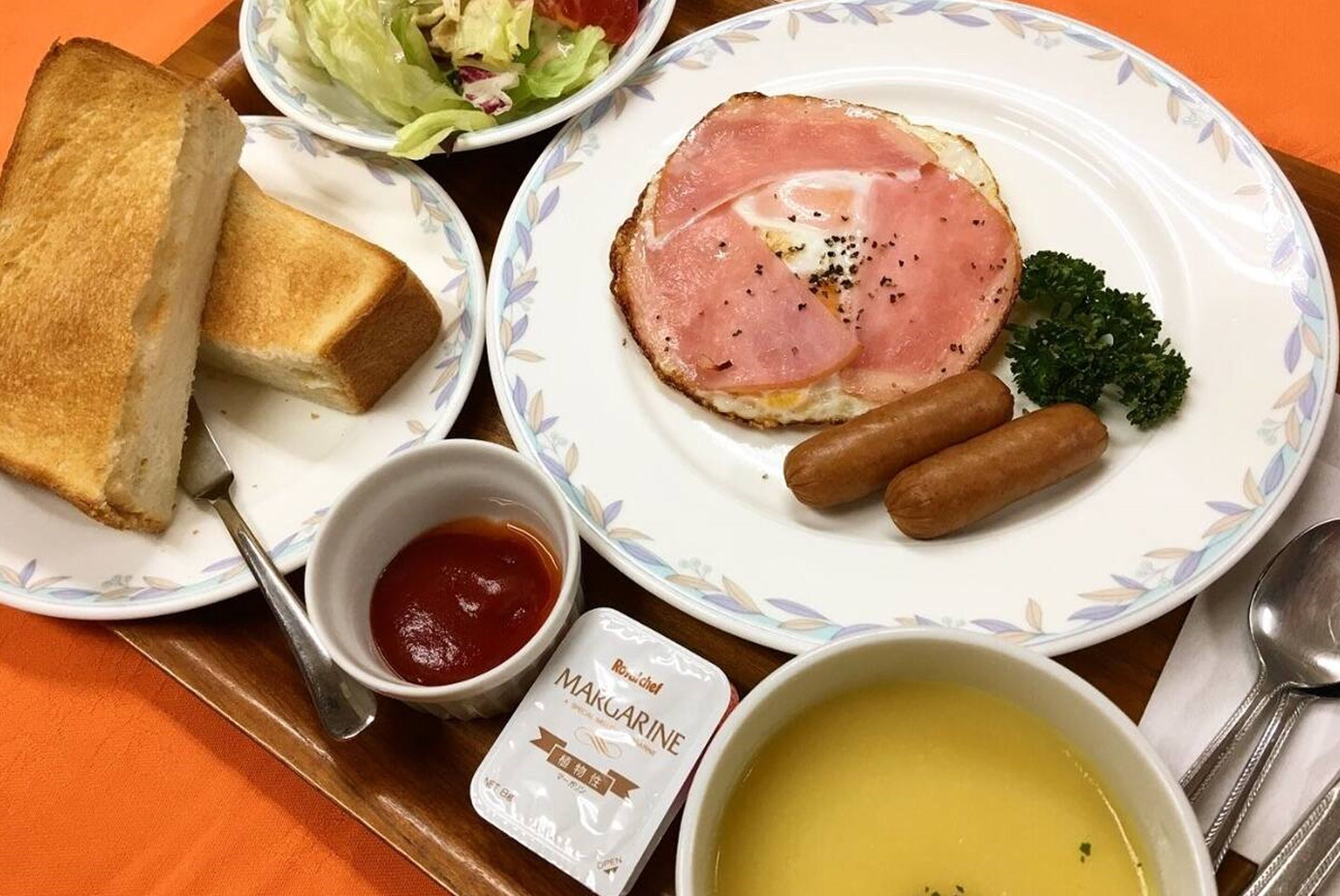 Bandeja de desayuno con tostadas, jamón, salchichas, un tazón de sopa, ensalada y una porción de margarina.