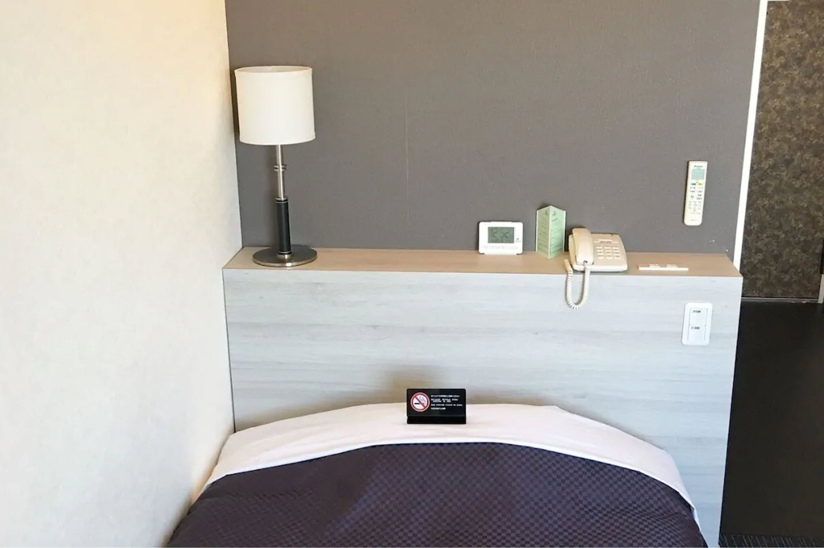 A hotel room with a bed, a bedside table with a lamp, a phone, a thermostat, and a sign indicating no smoking.