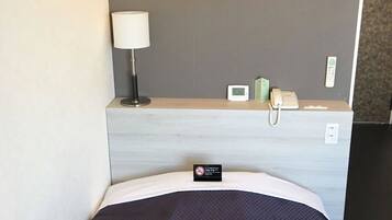 A hotel room with a bed, a bedside table with a lamp, a phone, a thermostat, and a sign indicating no smoking.