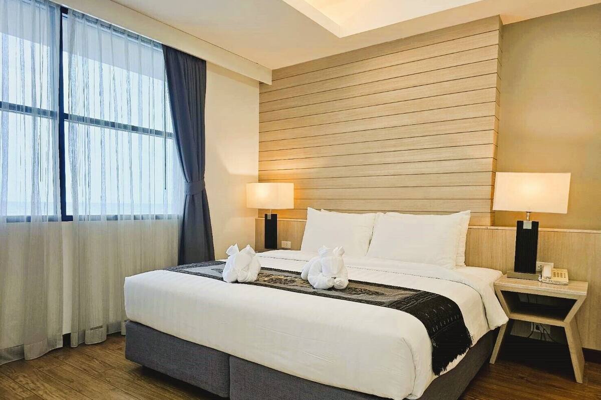 A hotel room with a large bed, two bedside lamps, a wooden headboard, and a window with sheer curtains.