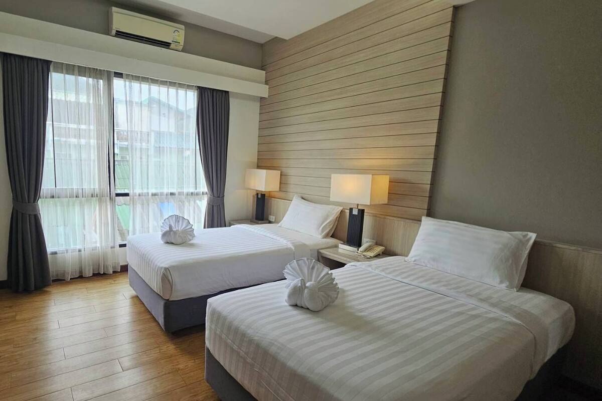 A hotel room with two beds, a wooden floor, a window with curtains, and a wall-mounted air conditioner.