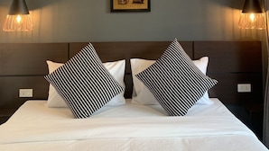 Une chambre d’hôtel moderne avec un grand lit, deux oreillers à rayures noires et blanches, un tableau encadré au mur et des lampes suspendues.