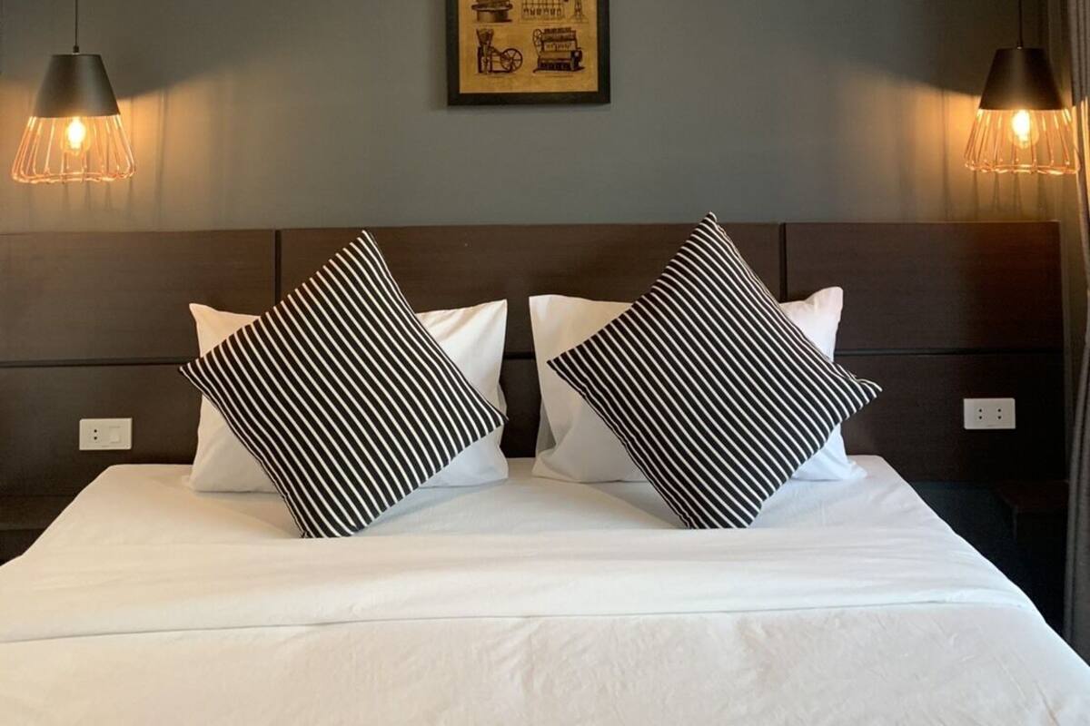 Une chambre d’hôtel moderne avec un grand lit, deux oreillers à rayures noires et blanches, un tableau encadré au mur et des lampes suspendues.
