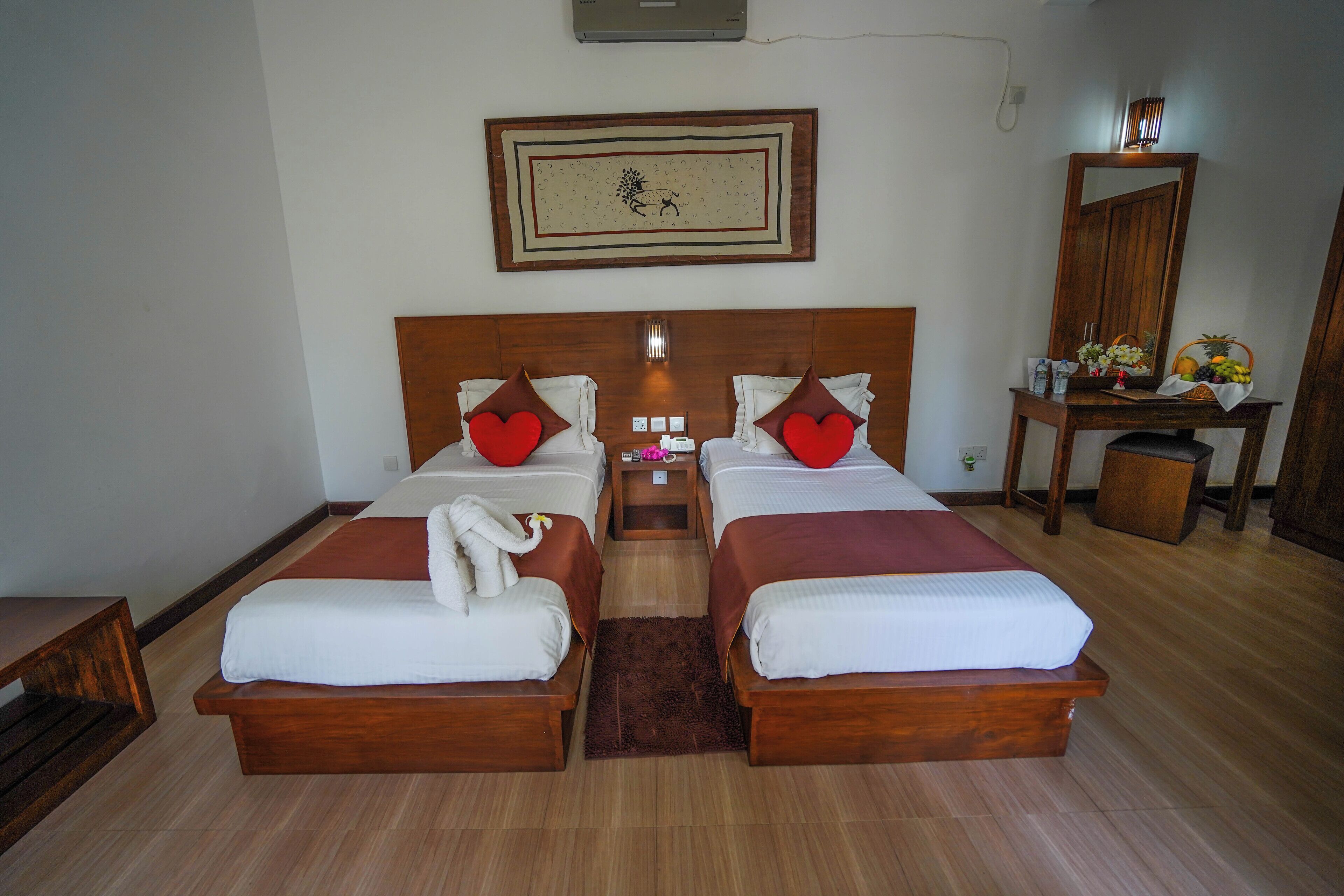 A double room with two beds, each with a wooden headboard, white bedding, and red pillows. There is a small table with a mirror and a fruit basket on the right side.