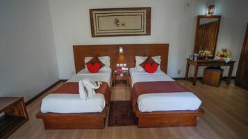 A double room with two beds, each with a wooden headboard, white bedding, and red pillows. There is a small table with a mirror and a fruit basket on the right side.