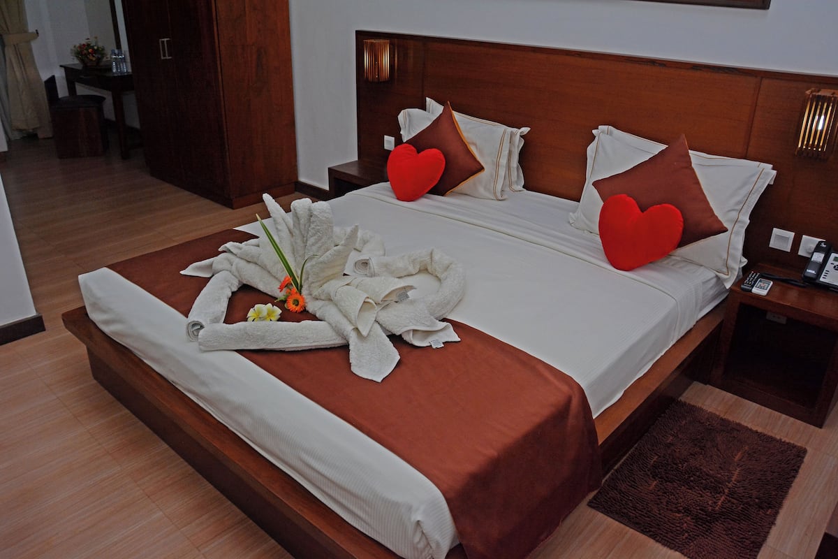 Una cama bien hecha con sábanas blancas, dos almohadas con forma de corazón rojo y un ramo de flores al lado.