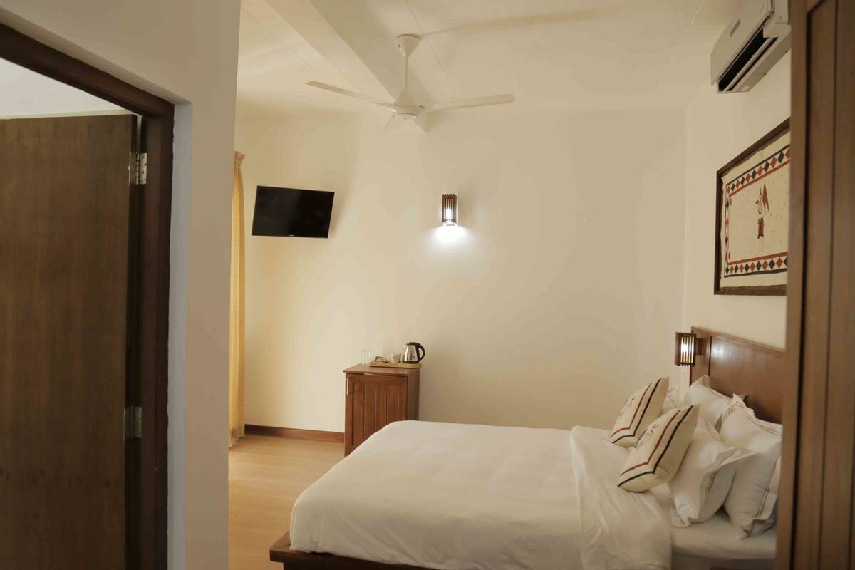 Un dormitorio con cama, ventilador de techo, mesita de noche con lámpara y televisor montado en la pared.