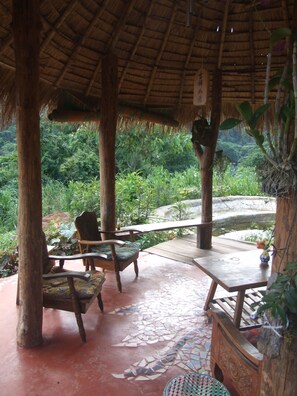 A covered outdoor seating area with wooden furniture and a thatched roof, surrounded by lush greenery. - Chiang Dao Roundhouses (Chiang Dao)