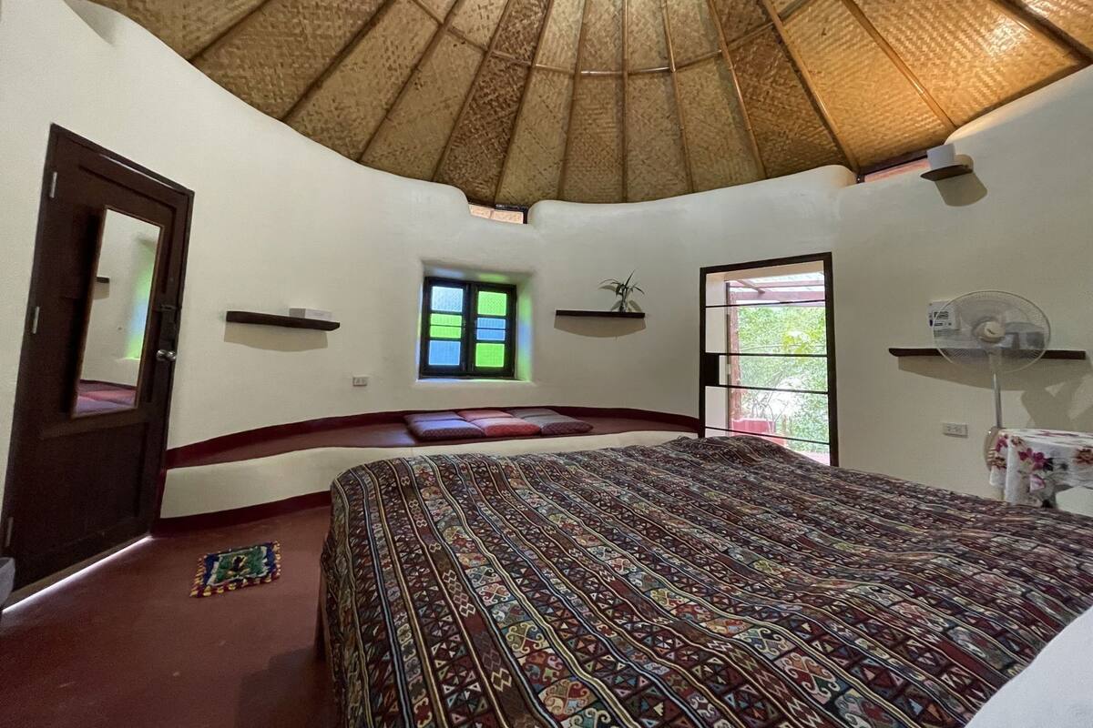 A room with a curved ceiling, a bed with patterned bedding, a small window, and a fan.