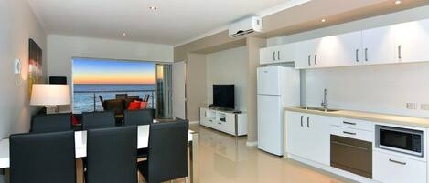 Una cocina moderna con armarios blancos, horno empotrado y microondas. Hay una zona de comedor con mesa y sillas, y un televisor sobre un mueble.