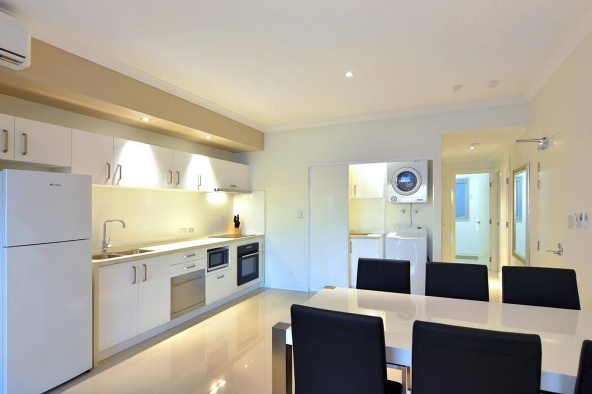 Una cocina moderna con armarios blancos, horno integrado y comedor con mesa blanca y sillas negras.