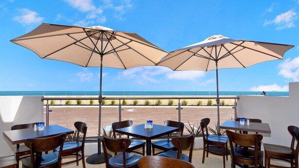 Une terrasse avec des tables et des chaises, deux grands parasols beiges et une vue sur l’océan.