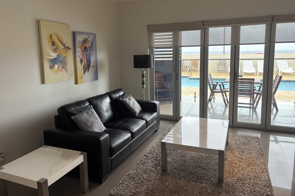 Una sala de estar con un sofá de cuero negro, una mesa de centro blanca y un amplio ventanal corredera que da a un área exterior con piscina y sillones de descanso.