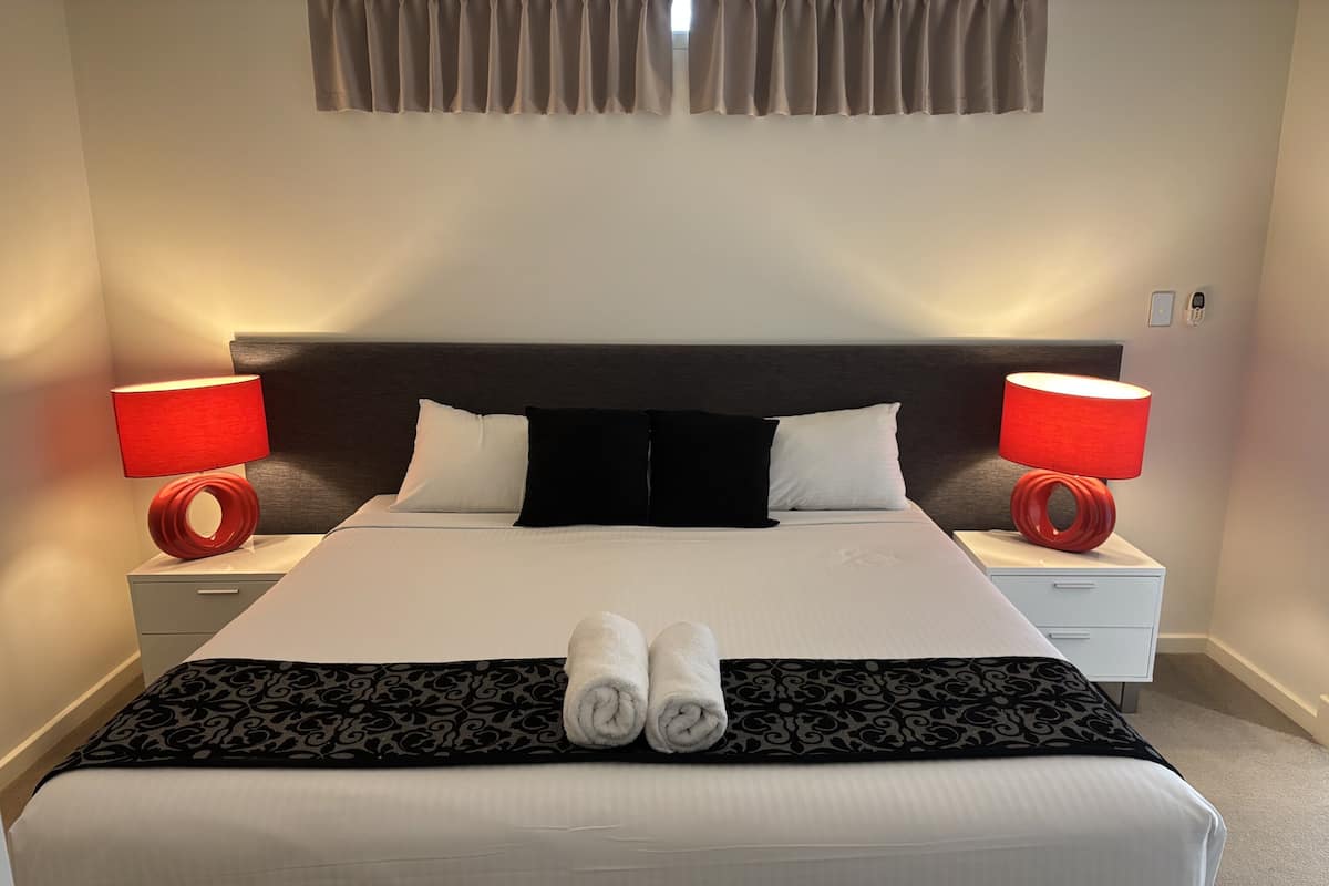 Una cama bien tendida con un cabecero negro, dos lámparas de noche y un par de toallas enrolladas sobre la cama.