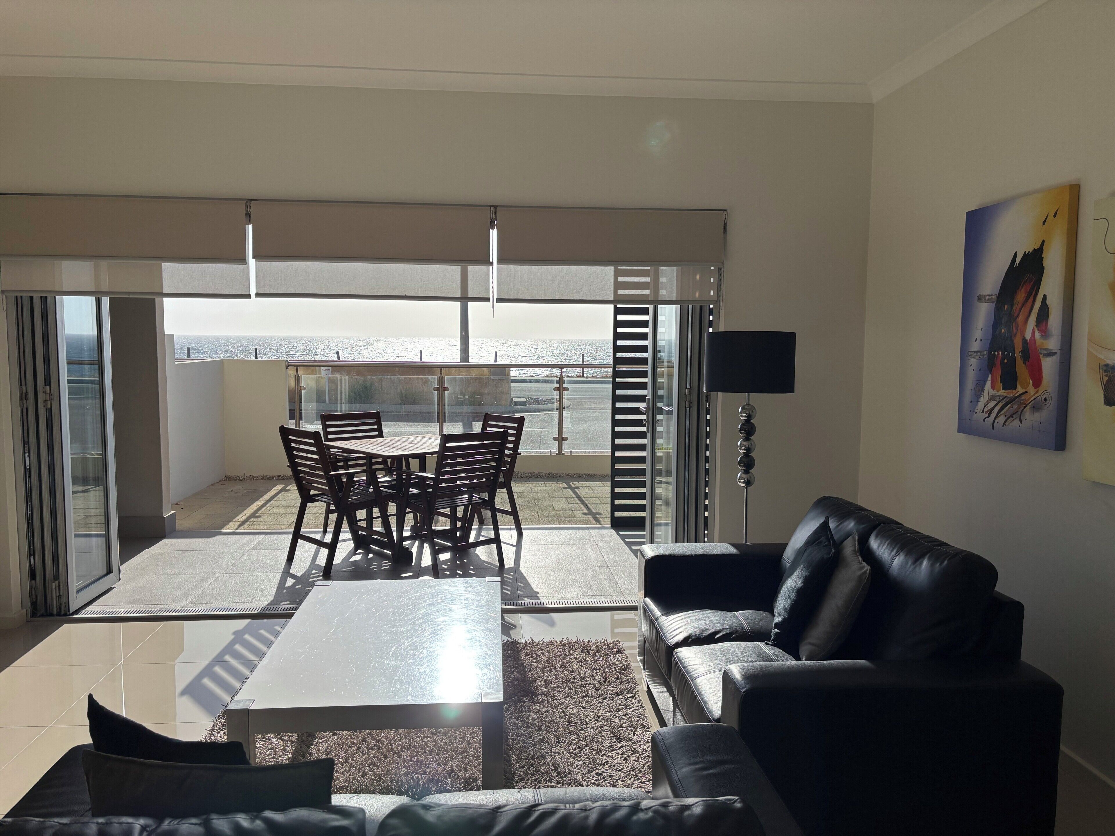 A modern living room with a glass door leading to a balcony, a black leather sofa, a coffee table, and a dining area with chairs.