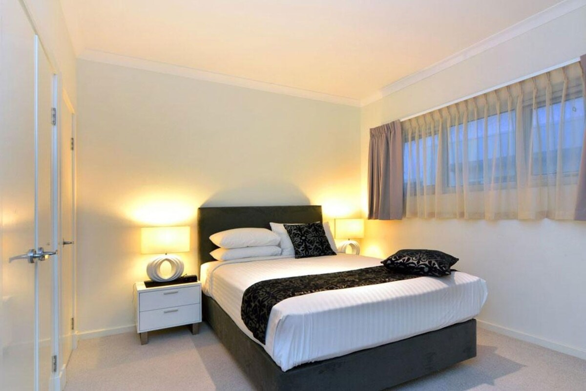 Een slaapkamer met een bed, nachtkastjes, een raam met gordijnen en een deur.