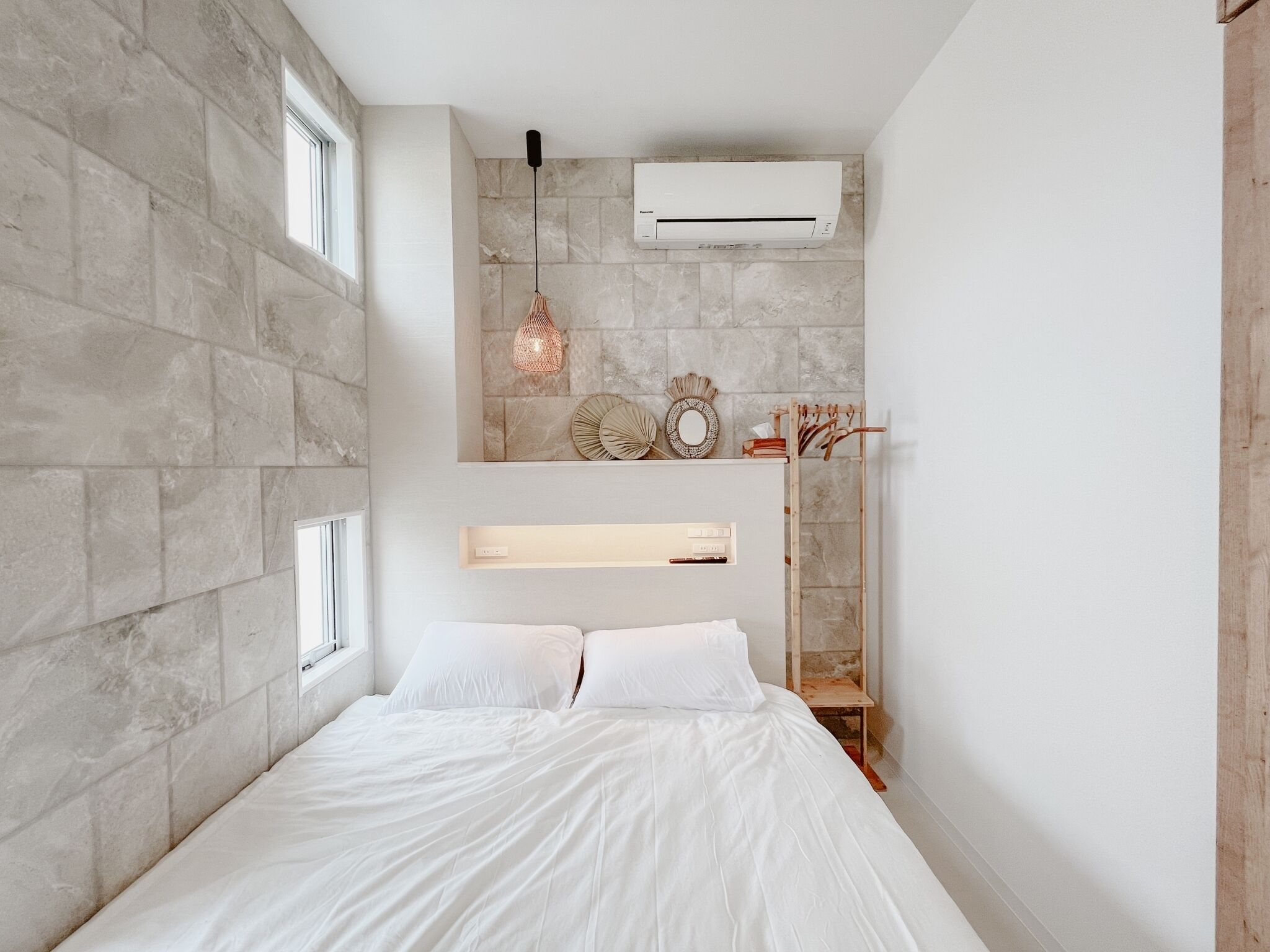A bedroom with a stone wall, a bed with white bedding, a hanging light fixture, and a wall-mounted air conditioner.
