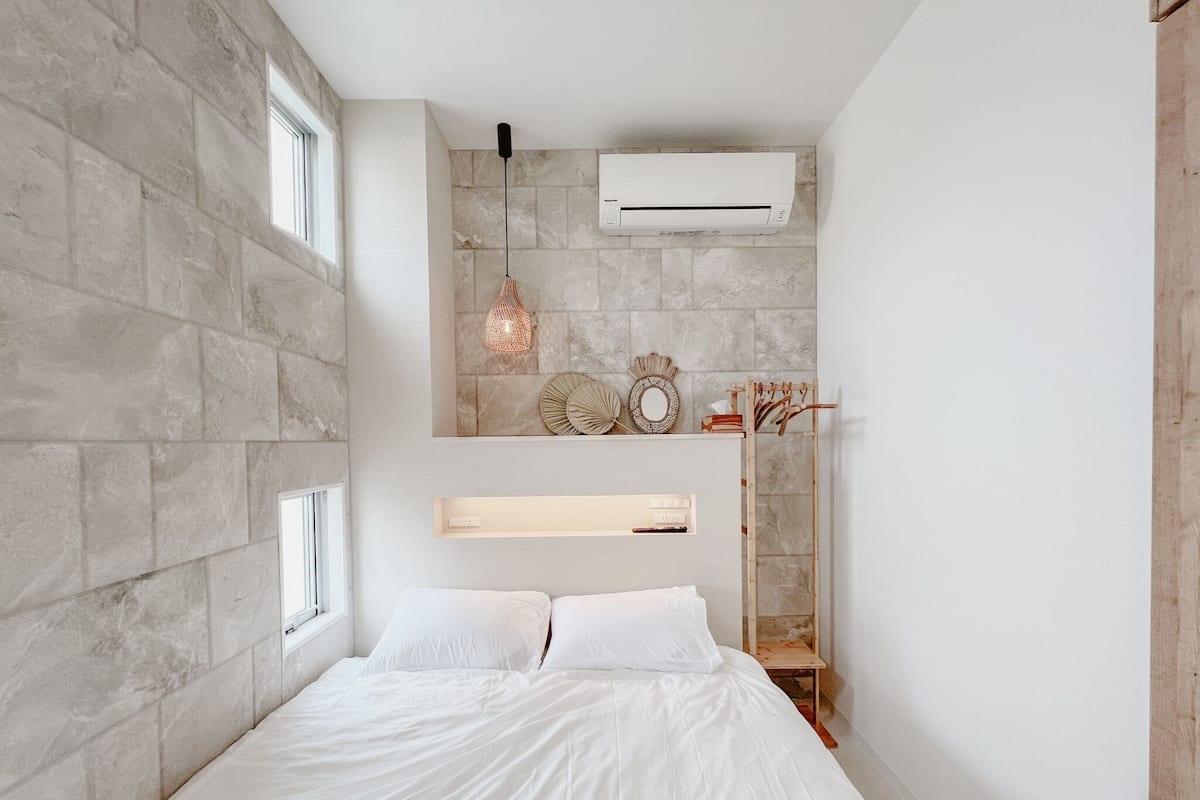 Un dormitorio con pared de piedra, cama con ropa blanca, lámpara colgante y aire acondicionado instalado.