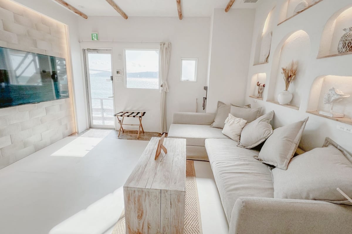 Una sala moderna con un televisor grande, una mesa de centro de madera y un sofá con cojines. Se ve una ventana con vistas al mar.