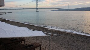 A beach with a large bridge in the background, wooden benches, and a white canopy. - BeachHouse Geragera (Kobe)