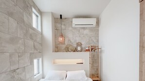 A bedroom with a stone wall, a bed with white bedding, a hanging light fixture, and a mounted air conditioner. - BeachHouse Geragera (Kobe)