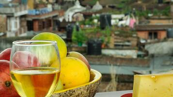 Una mesa con una copa de vino, un bol de fruta y un plato de queso, frente a un paisaje urbano con arquitectura tradicional y montañas.