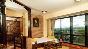 Una habitación con una escalera de madera, pared de ladrillo, cama con almohada amarilla, mesa con lámpara y un ventanal con vistas a la ciudad.