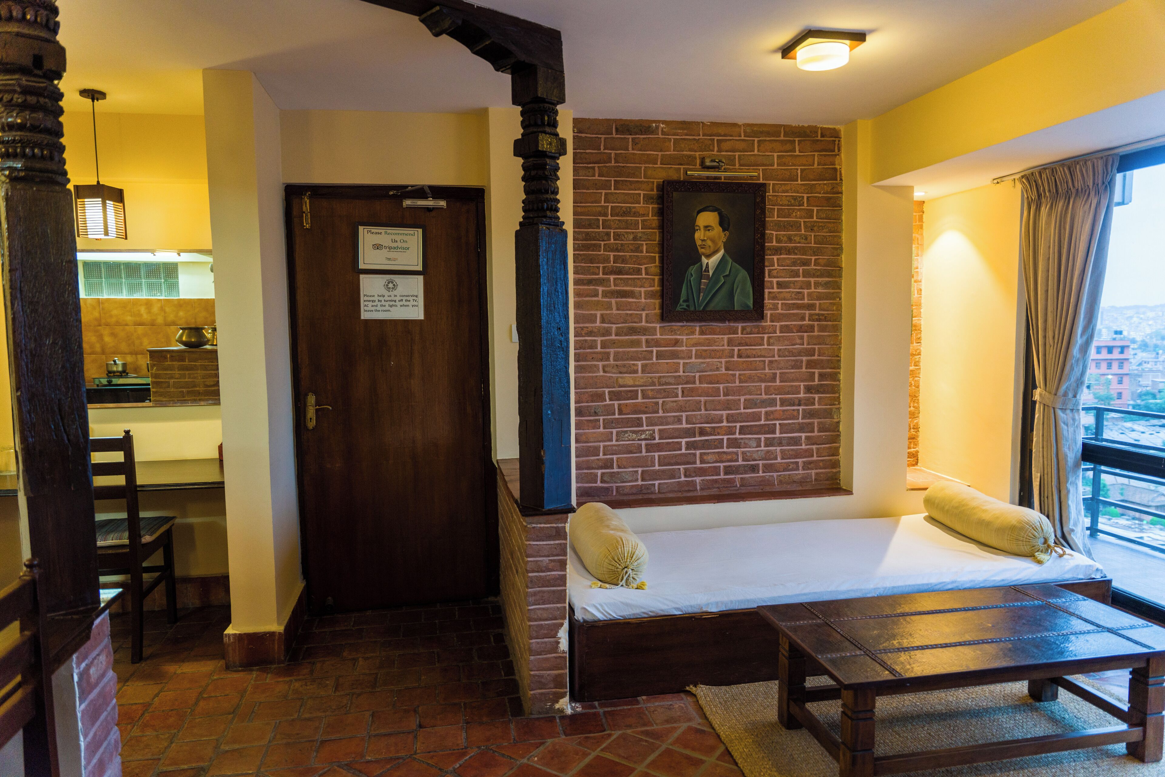 Une pièce comprenant un lit, un banc en bois, un coin repas avec une table et des chaises, et un mur en briques orné d’un portrait encadré.