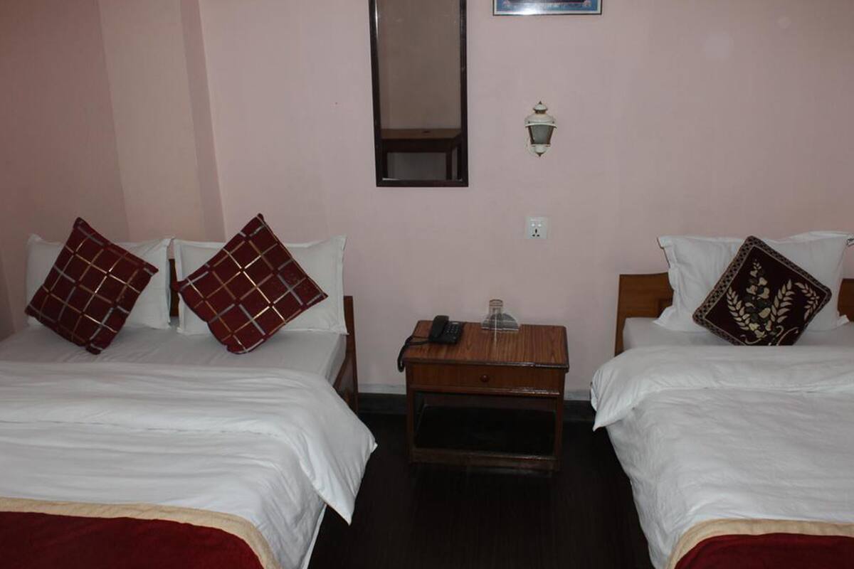 Quarto de hotel com duas camas, cada uma com roupa de cama branca e almofadas decorativas. Há uma mesa de cabeceira de madeira entre as camas, com uma lâmpada e um copo sobre ela.
