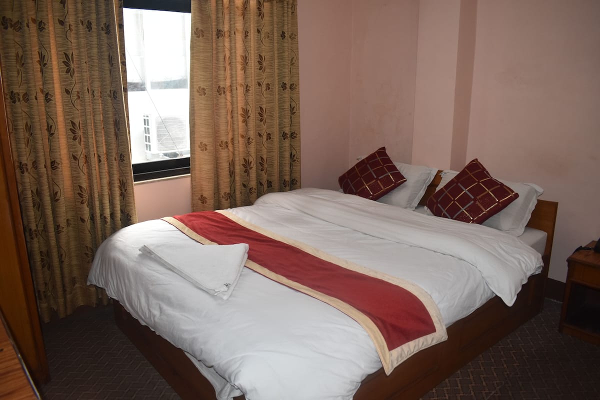 Cama bem arrumada com roupa de cama branca e vermelha, uma janela com cortinas e uma mesinha de cabeceira de madeira.