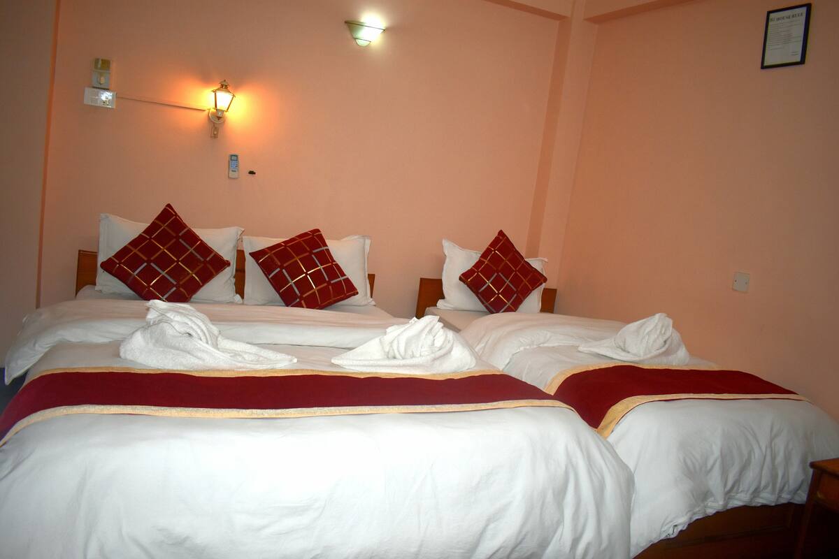 Quarto de hotel com duas camas, cada uma com lençóis brancos e almofadas padronadas em vermelho e branco. Há duas luminárias fixadas na parede e um aviso emoldurado.