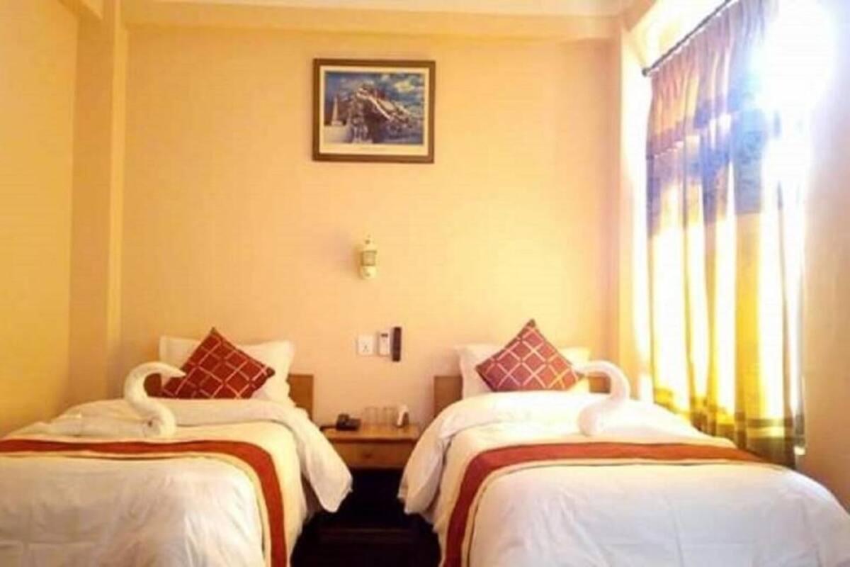 Quarto de hotel com duas camas, cada uma com roupa de cama branca e detalhes vermelhos, uma mesa de cabeceira, um quadro na parede e uma janela com cortinas.