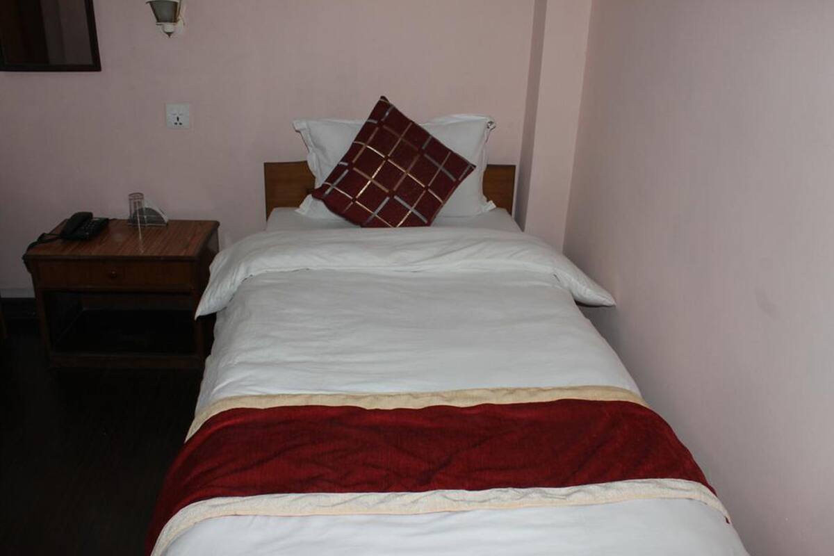 Uma cama de solteiro com colcha branca e vermelha, uma mesinha de cabeceira de madeira com telefone e abajur, e uma parede lisa com luminária.