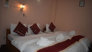 A hotel room with two beds, each with white linens and red pillows. There is a red throw blanket on one bed, and towels are neatly arranged on the other. A wall-mounted lamp provides lighting. - Festoon Hotel (Kathmandu)