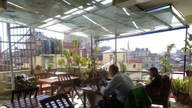 A covered outdoor seating area with wooden tables and chairs, potted plants, and a view of the cityscape. - Festoon Hotel (Kathmandu)