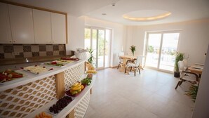 A bright, spacious room with a buffet table filled with fruits and cheeses, a dining area with chairs, and large windows offering a view of the outdoors. - Hotel Aysima (Kas)