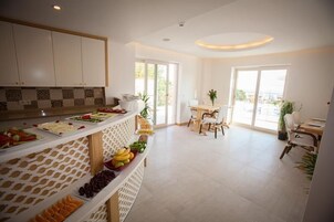Una habitación luminosa y amplia con una mesa de buffet repleta de frutas y quesos, un comedor con sillas y ventanas grandes que ofrecen vistas al exterior.