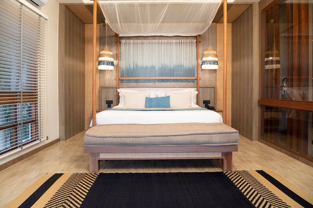 Une chambre d’hôtel comprenant un lit, des tables de chevet, une fenêtre avec des stores, un rideau et un luminaire suspendu.