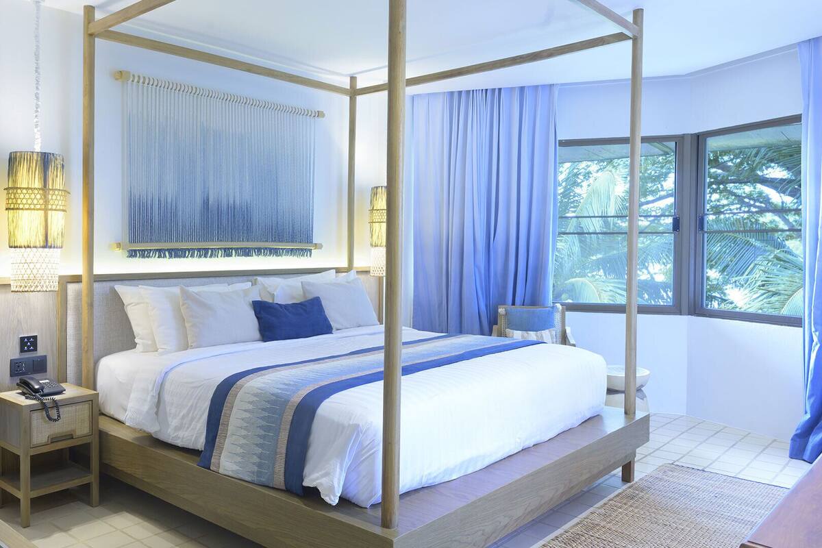 Un lit à baldaquin avec du linge de lit blanc et des touches de bleu, une table de chevet avec une lampe, et une vue sur les palmiers par une grande fenêtre.