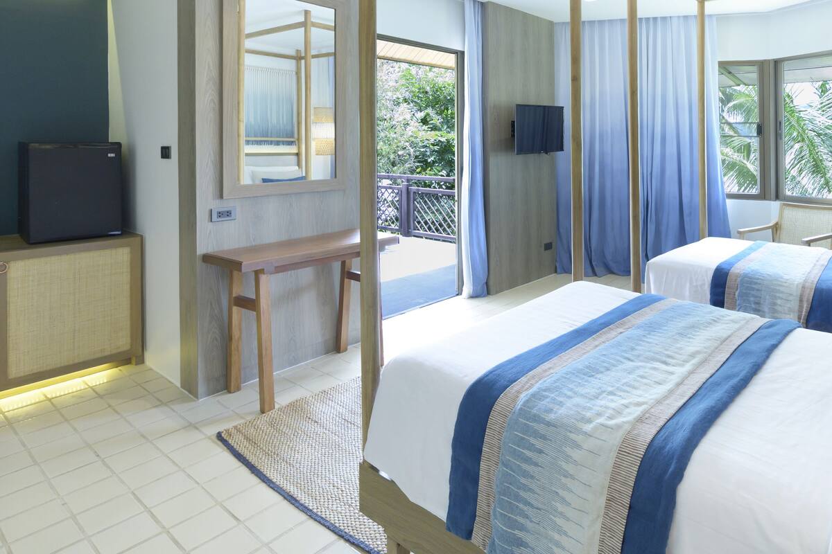 Une chambre d’hôtel avec deux lits, une télévision, un bureau et un balcon offrant une vue sur les palmiers.