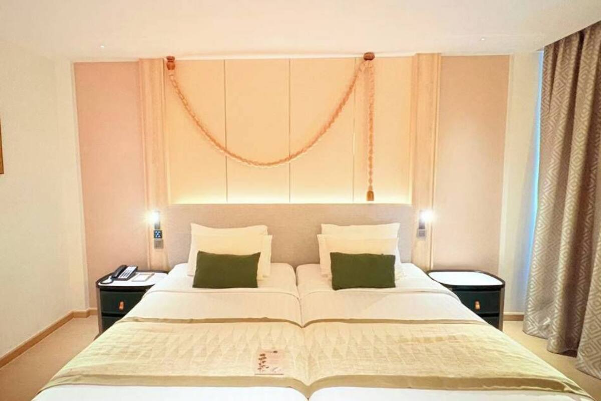 Un lit double avec une couvre-lit beige et deux oreillers, une table de chevet avec un téléphone et un luminaire fixé au mur.