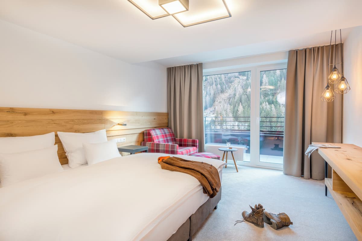 A hotel room with a large bed, a wooden headboard, a plaid armchair, a small table, and a view of a snowy landscape through the window.