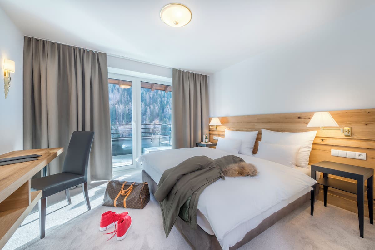 A hotel room with a bed, a desk, a chair, a suitcase, and a view of the outdoors.