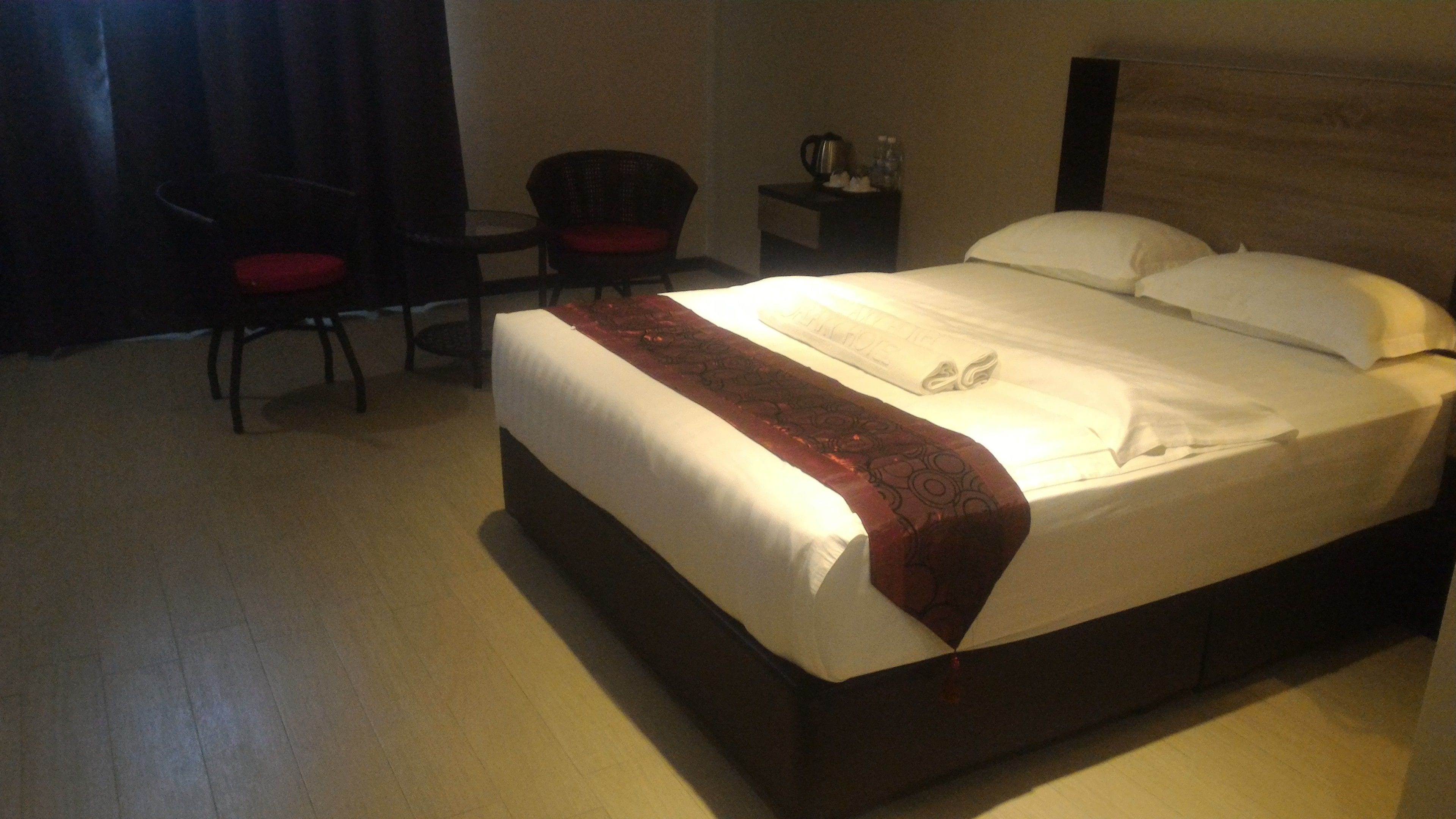 A hotel room with a large bed, a chair, a small table, and a nightstand with a lamp.